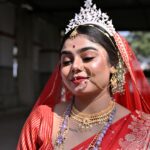 Elegant Bengali bride in red saree with intricate jewelry and makeup outdoors in Purulia.